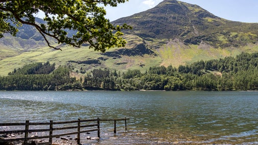 Lake with a partially submerged fence in the foreground, looking across to a sunlit green mountain and woodland beneath a clear blue sky.
